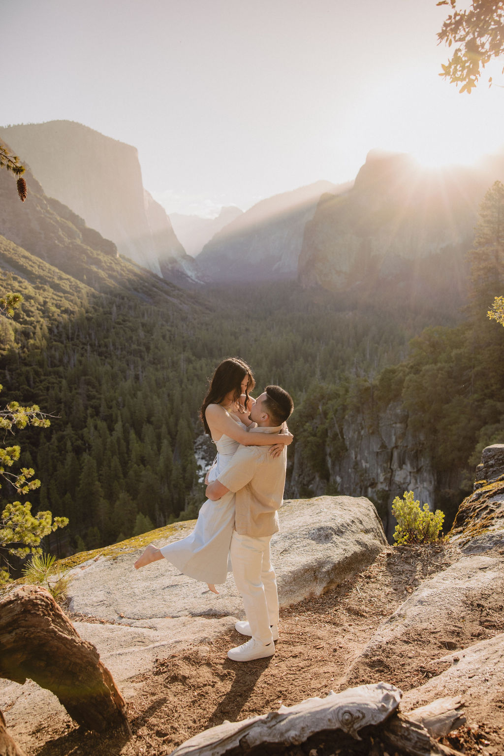 A couple embraces on a rocky ledge overlooking a forested valley with mountains in the background at sunset. | How to Plan a Surprise Proposal at Yosemite National Park