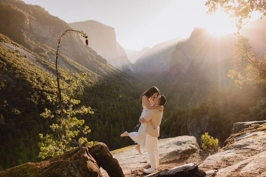 A couple embraces on a rocky ledge overlooking a forested valley with mountains in the background at sunset. | How to Plan a Surprise Proposal at Yosemite National Park