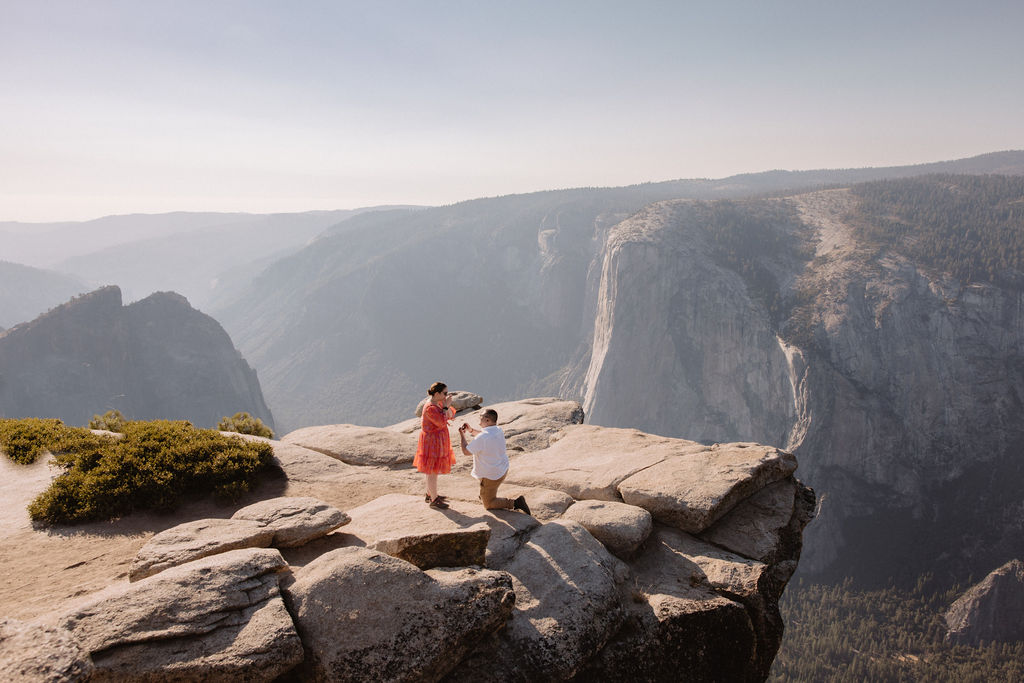 A person in a red dress and a person in a suit stand on a rocky cliff, with a vast mountain landscape in the background under a clear sky.