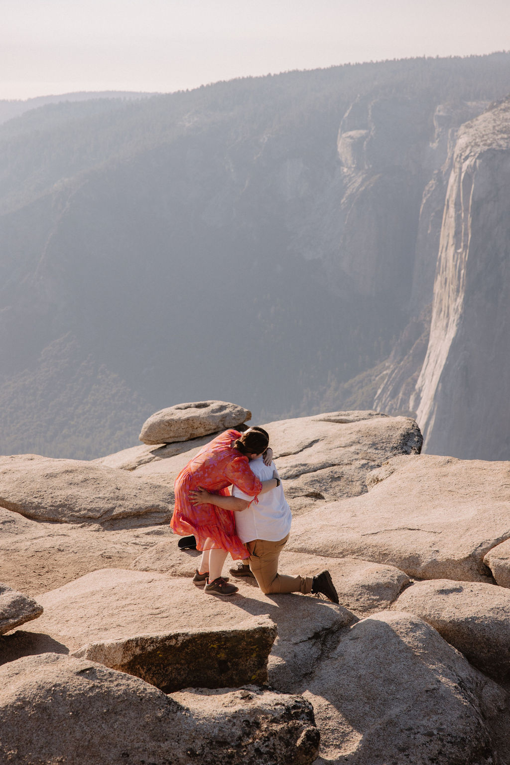 A couple embraces on a rocky cliff with a vast canyon in the background under a clear sky for a surprise proposal at Yosemite 