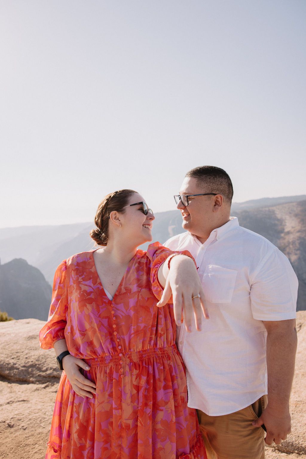 A couple stands on a rocky overlook with a mountainous landscape in the background. The woman wears a bright patterned dress, and the man wears a white shirt and glasses for a surprise proposal proposal at yosemite