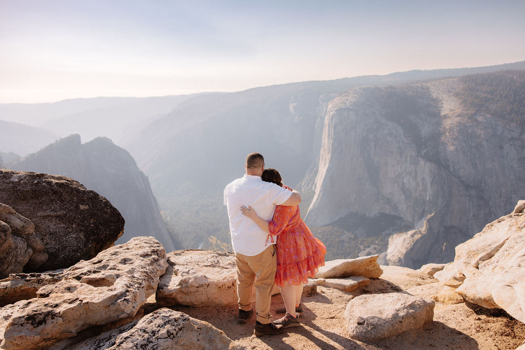 A couple stands on a rocky cliff, embracing and gazing at a vast mountain vista under a clear sky for a surprise proposal at Yosemite national park