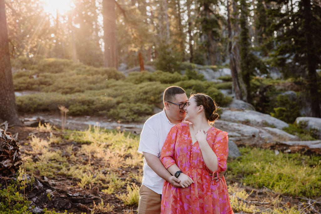 A couple embraces in a sunlit forest, surrounded by tall trees and greenery.