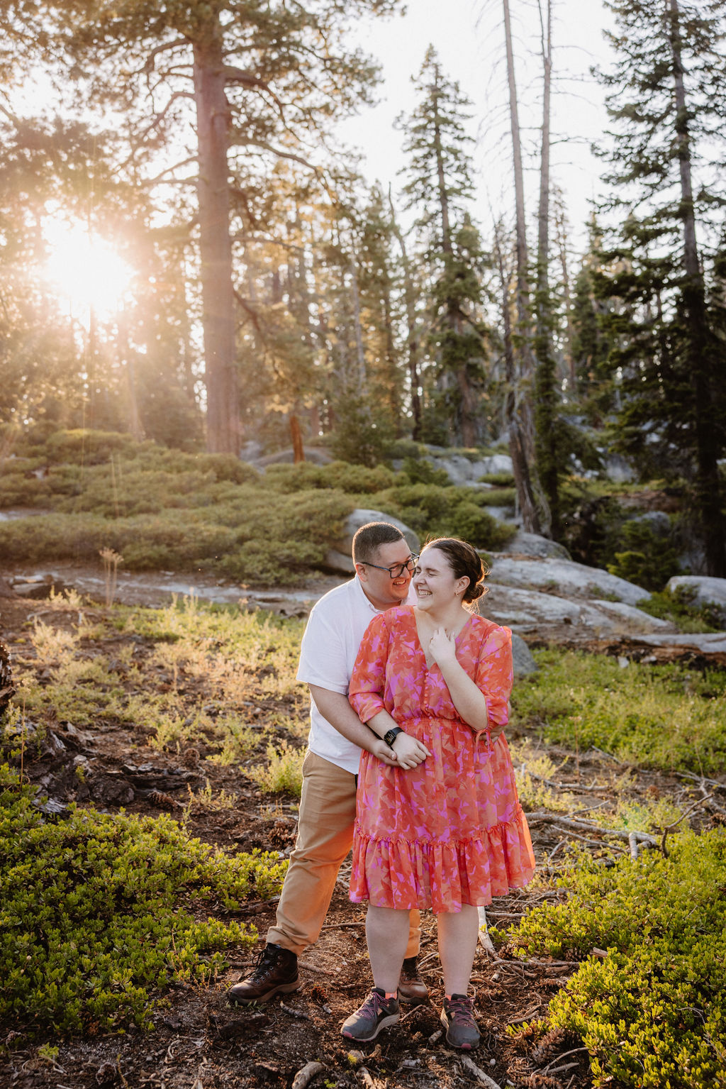 A couple embraces in a sunlit forest, surrounded by tall trees and greenery.