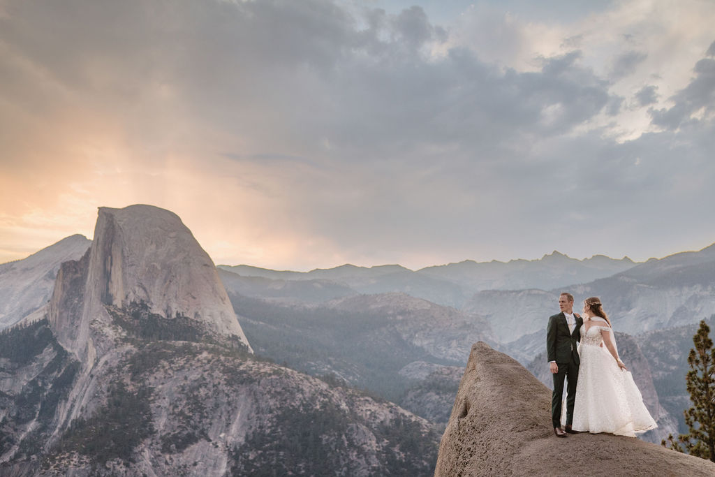 A couple in wedding attire stands on a rocky ledge, overlooking a mountainous landscape at sunrise at an elopement at Yosemite National park in glacier point and mariposa grove