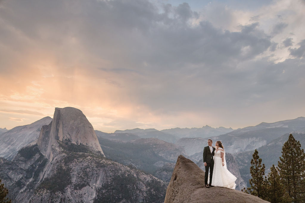 A couple in wedding attire stands on a rocky ledge, overlooking a mountainous landscape at sunrise at an elopement at Yosemite National park in glacier point and mariposa grove