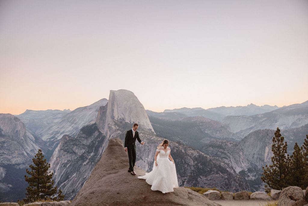 A couple in wedding attire stands on a rocky ledge, overlooking a mountainous landscape at sunrise at an elopement at Yosemite National park 