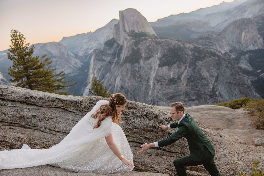 A couple in wedding attire stands on a rocky ledge, overlooking a mountainous landscape at sunrise at an elopement at Yosemite National park in glacier point and mariposa grove