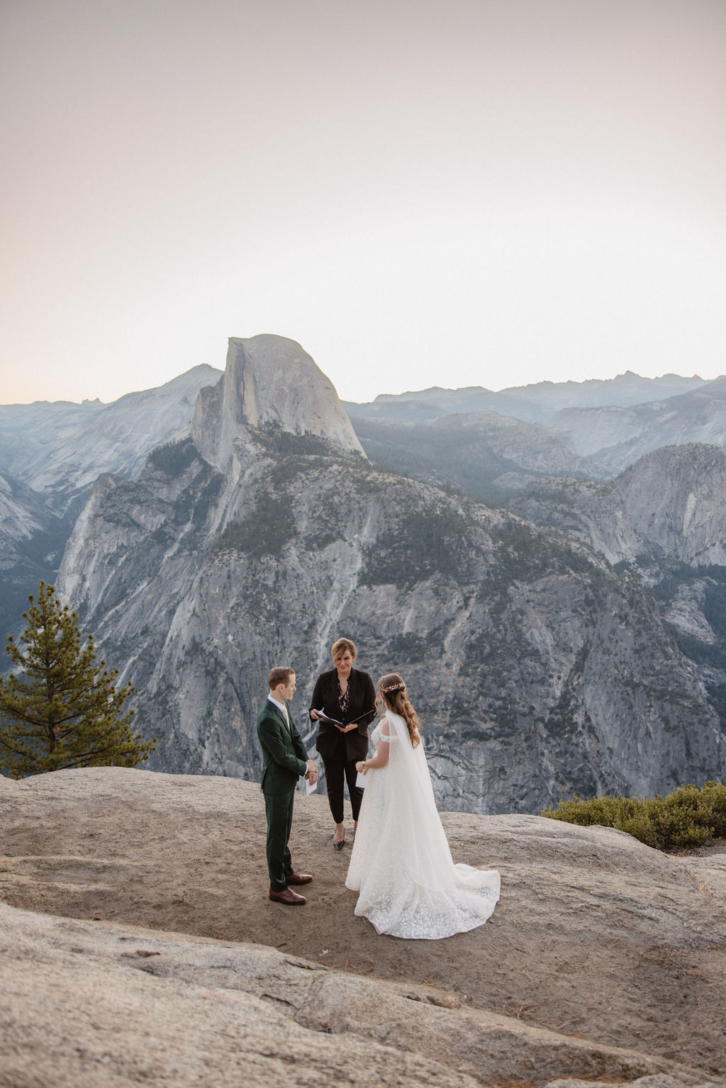 A couple exchanges vows with an officiant on a rocky overlook, with a mountain landscape in the background.