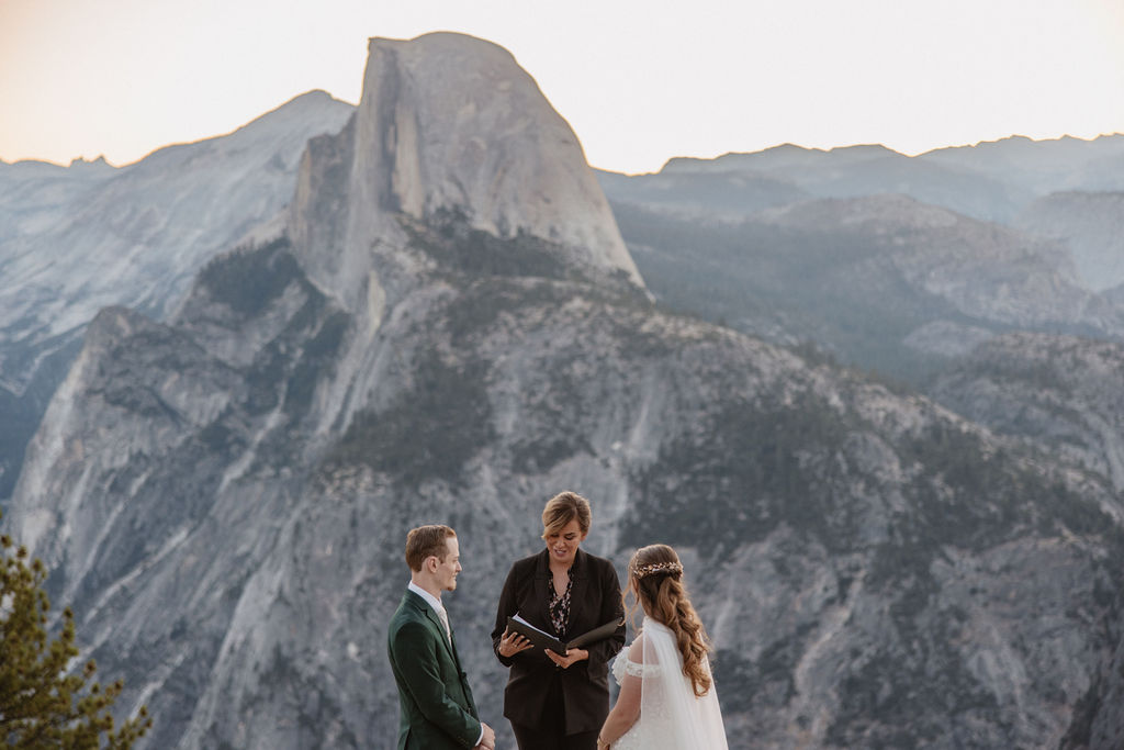 A couple exchanges vows with an officiant on a rocky overlook, with a mountain landscape in the background for a Yosemite elopement at Glacier Point and Mariposa Grove