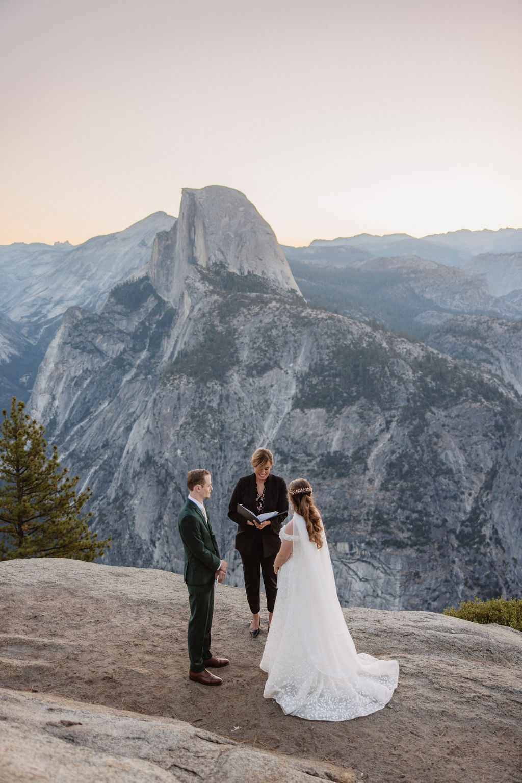 A couple exchanges vows with an officiant on a rocky overlook, with a mountain landscape in the background for a Yosemite elopement at Glacier Point and Mariposa Grove