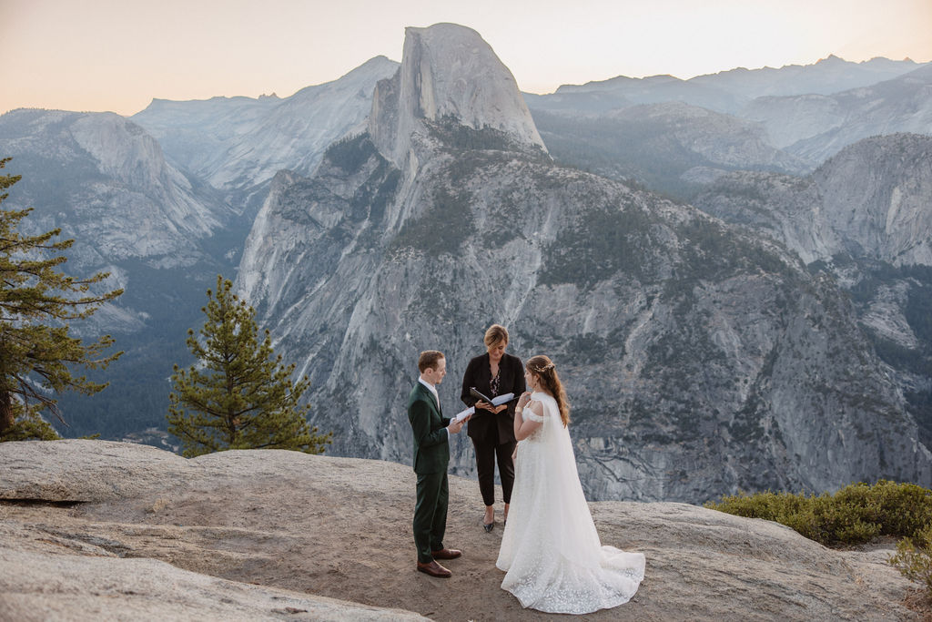 A couple exchanges vows with an officiant on a rocky overlook, with a mountain landscape in the background for a Yosemite elopement at Glacier Point and Mariposa Grove