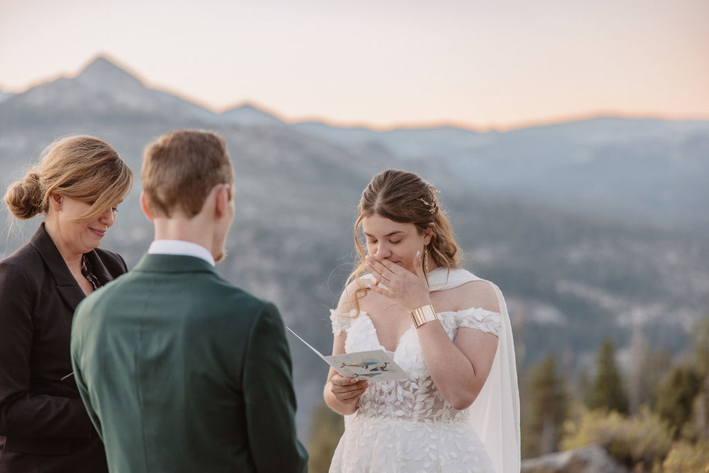 A couple exchanges vows with an officiant on a rocky overlook, with a mountain landscape in the background.