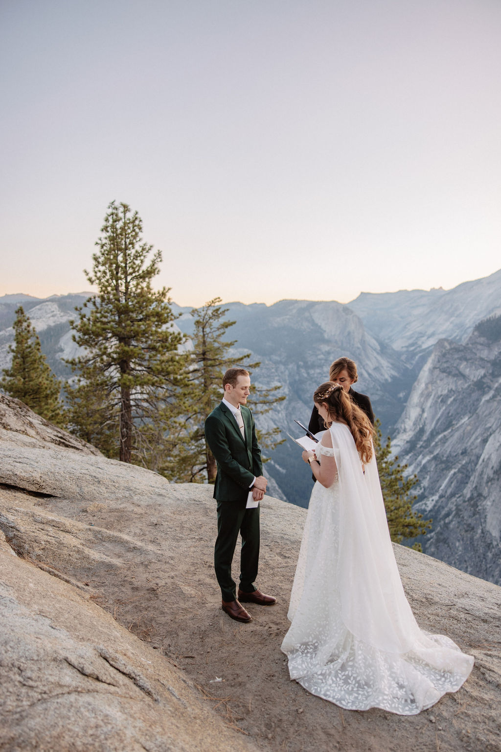 A couple exchanges vows with an officiant on a rocky overlook, with a mountain landscape in the background for a Yosemite elopement at Glacier Point and Mariposa Grove