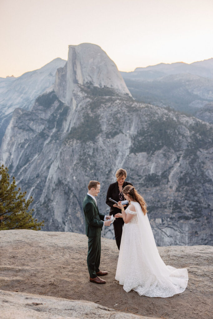 A couple exchanges vows with an officiant on a rocky overlook, with a mountain landscape in the background for a Yosemite elopement at Glacier Point and Mariposa Grove