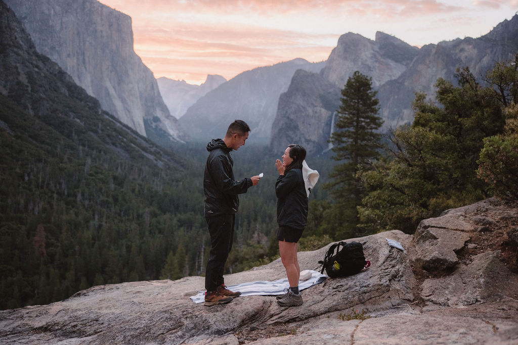 Two people stand facing each other on a rocky ledge with a scenic mountain and forest landscape in the background. One of them appears to be holding a small item. | How to Plan a Surprise Proposal at Yosemite National Park