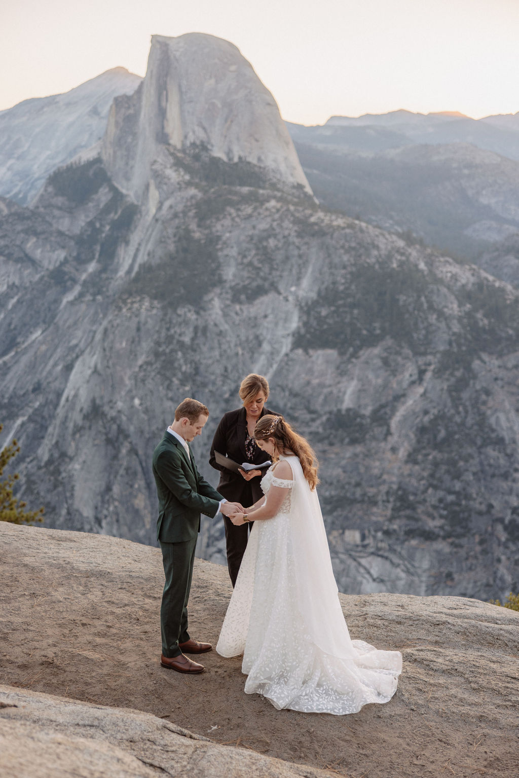 A couple exchanges vows with an officiant on a rocky overlook, with a mountain landscape in the background for a Yosemite elopement at Glacier Point and Mariposa Grove