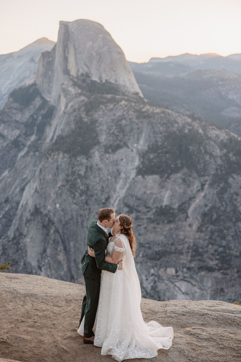 A couple exchanges vows with an officiant on a rocky overlook, with a mountain landscape in the background.