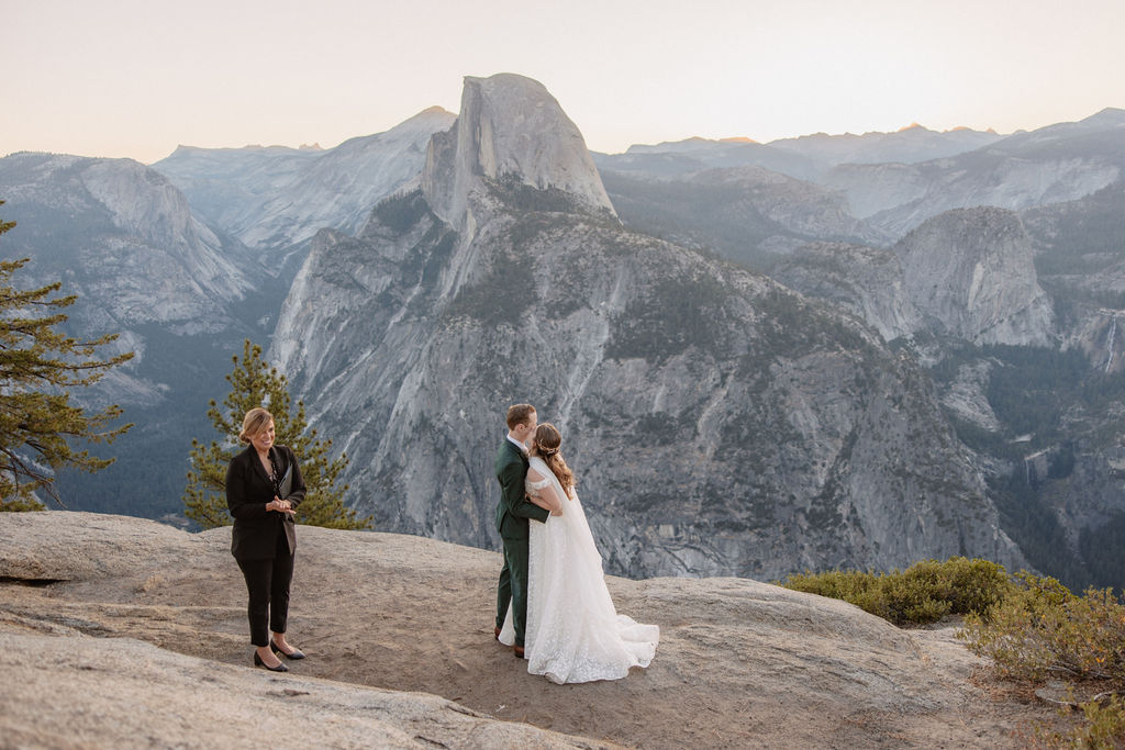 A couple exchanges vows with an officiant on a rocky overlook, with a mountain landscape in the background for a Yosemite elopement at Glacier Point and Mariposa Grove