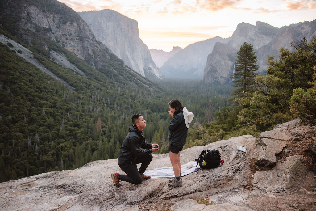 A person kneels proposing to another person on a rocky overlook with a forested valley and mountains in the background. A backpack and blanket are nearby. How to Plan a Surprise Proposal at Yosemite National Park