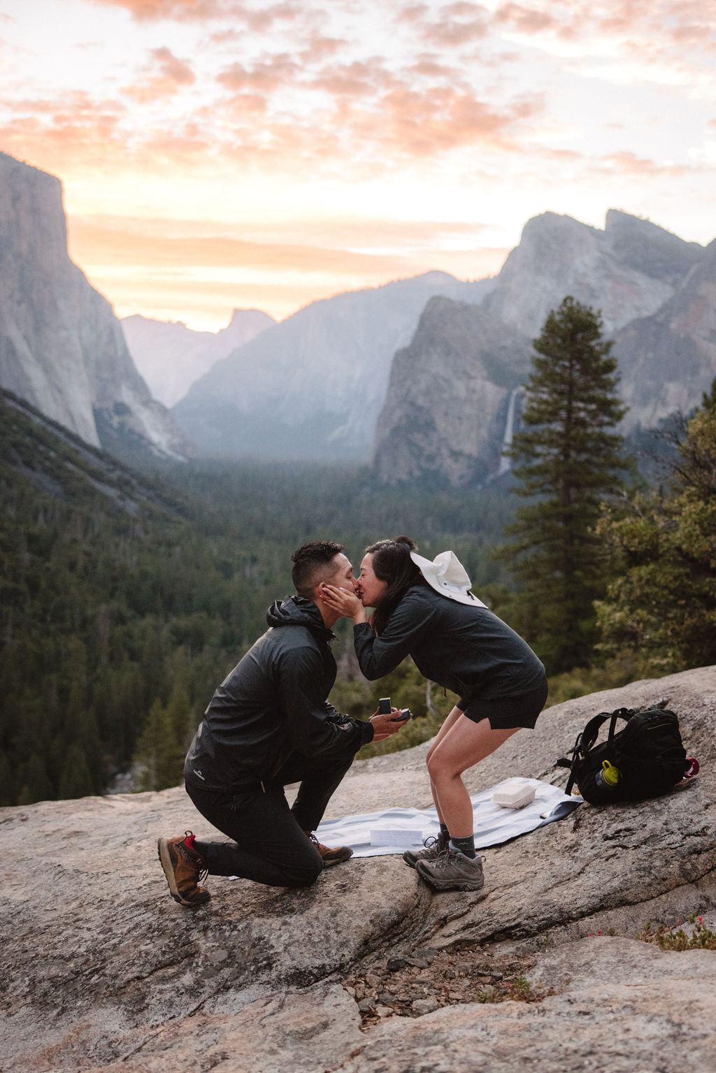 A couple kisses while kneeling on a rocky ledge, with a view of a forested valley and mountains in the background at sunset.