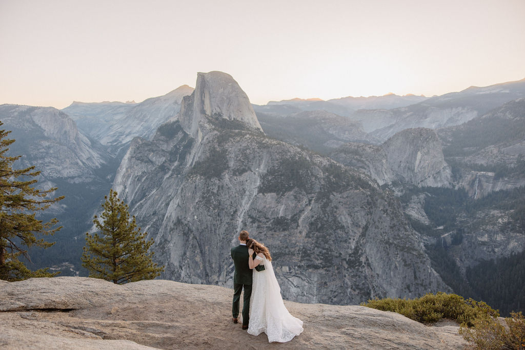 A couple in wedding attire stands on a rocky ledge, overlooking a mountainous landscape at sunrise at an elopement at Yosemite National park 