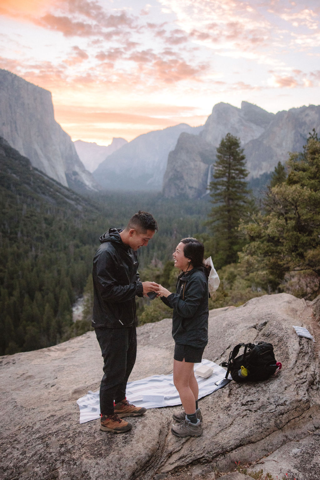 Two people stand on a rocky ledge at sunset in a mountainous landscape, interacting with each other. A backpack and blanket are on the ground nearby.