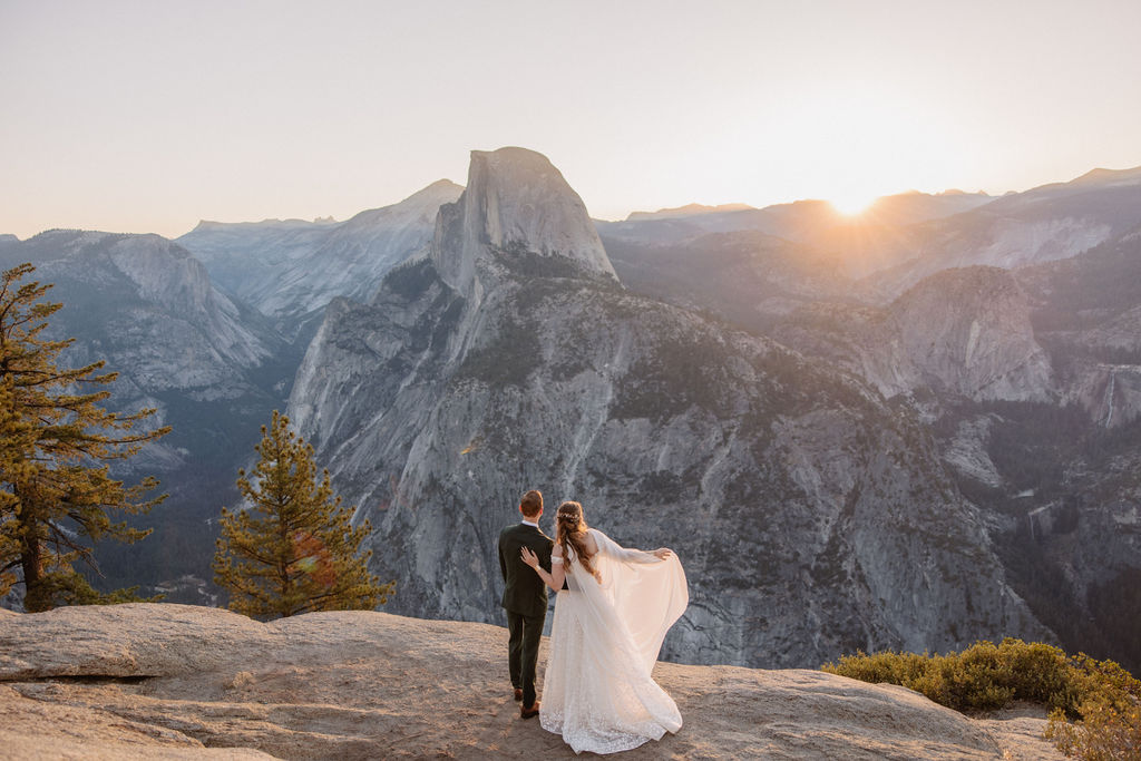 A couple in wedding attire stands on a rocky ledge, overlooking a mountainous landscape at sunrise at an elopement at Yosemite National park 