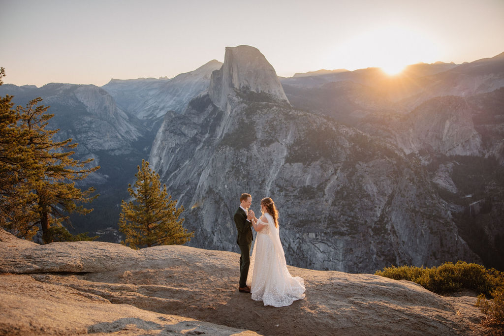 A couple in wedding attire stands on a rocky ledge, overlooking a mountainous landscape at sunrise at an elopement at Yosemite National park 