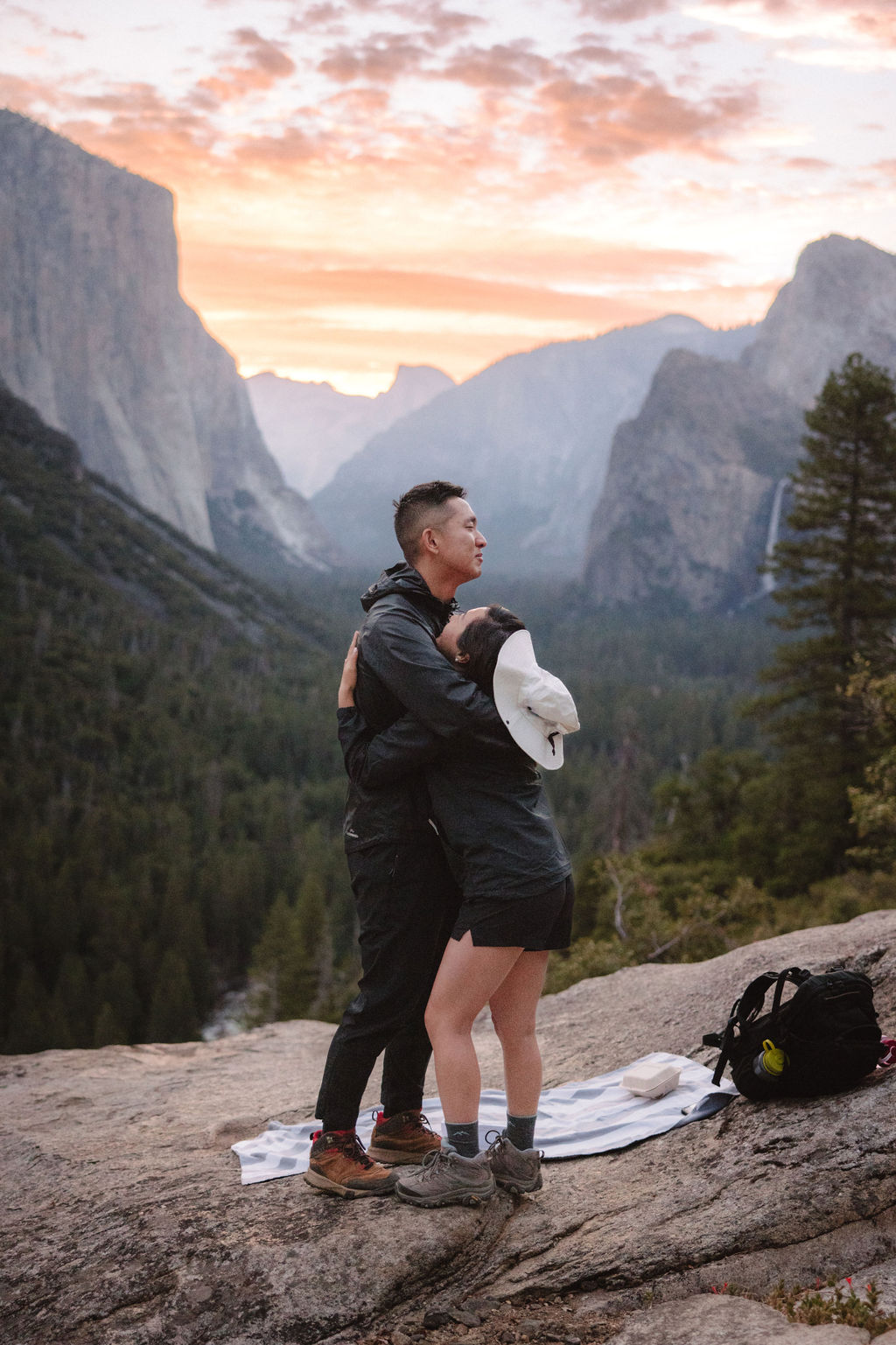 A couple hugs on a scenic overlook with mountains and a sunset in the background. A backpack and blanket are on the ground nearby.