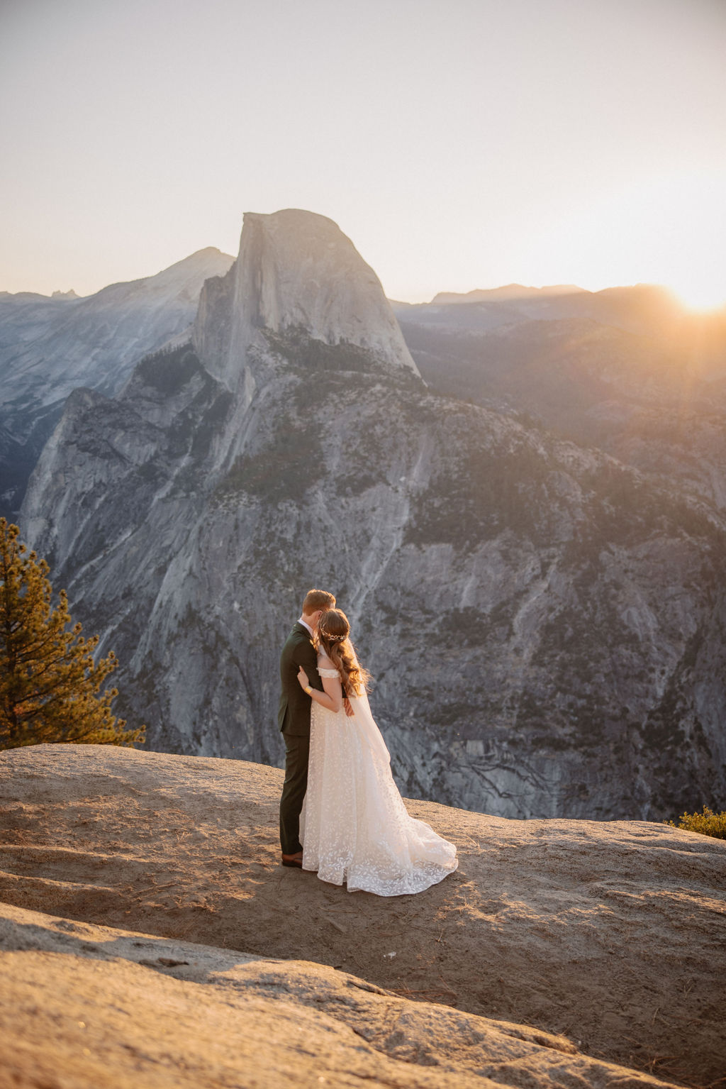 A couple in wedding attire stands on a rocky ledge, overlooking a mountainous landscape at sunrise at an elopement at Yosemite National park in glacier point and mariposa grove