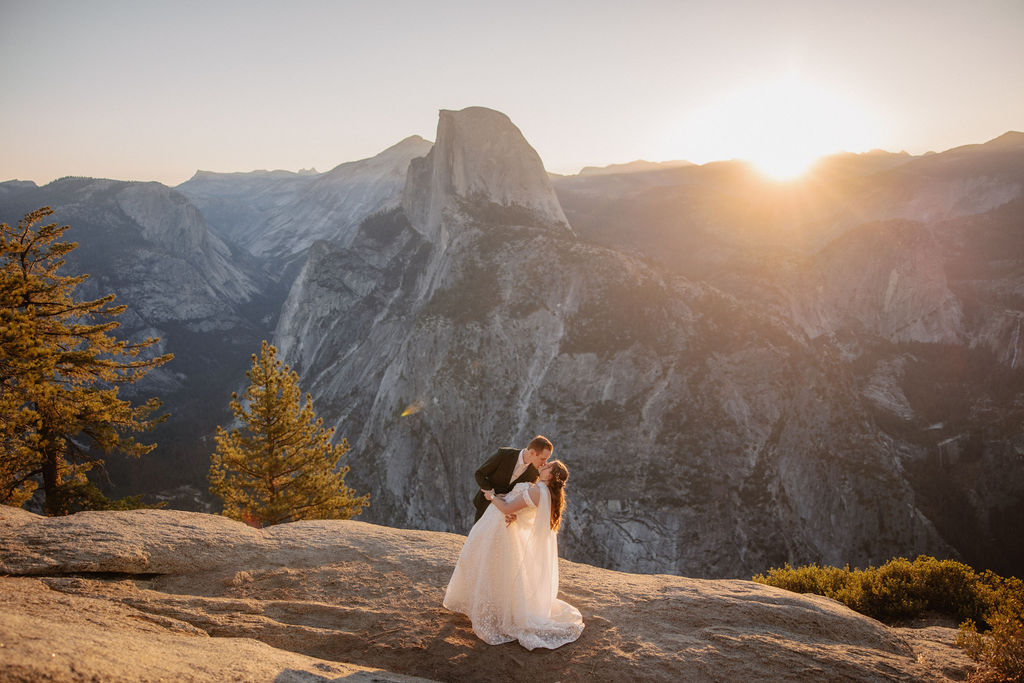 A couple in wedding attire stands on a rocky ledge, overlooking a mountainous landscape at sunrise at an elopement at Yosemite National park in glacier point and mariposa grove