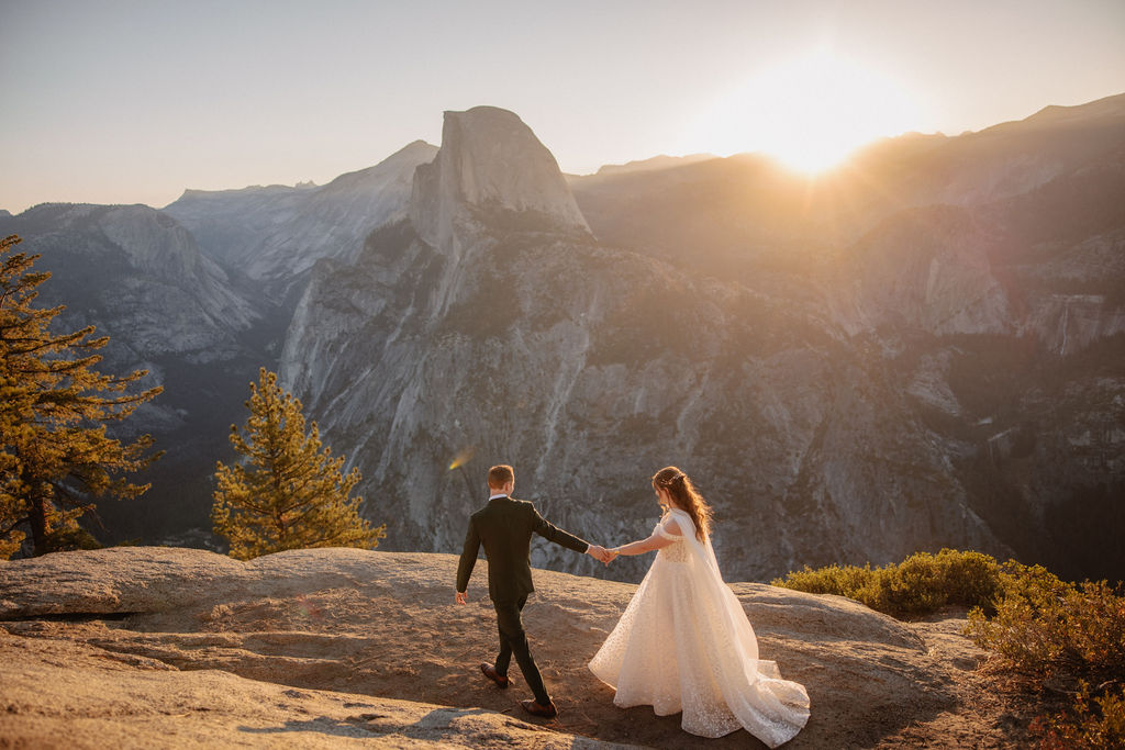 A couple in wedding attire stands on a rocky ledge, overlooking a mountainous landscape at sunrise at an elopement at Yosemite National park in glacier point and mariposa grove