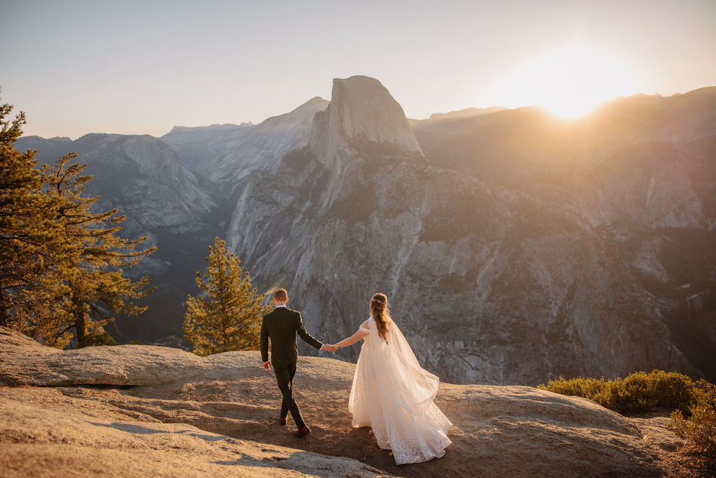 A couple in wedding attire stands on a rocky ledge, overlooking a mountainous landscape at sunrise at an elopement at Yosemite National park in glacier point and mariposa grove