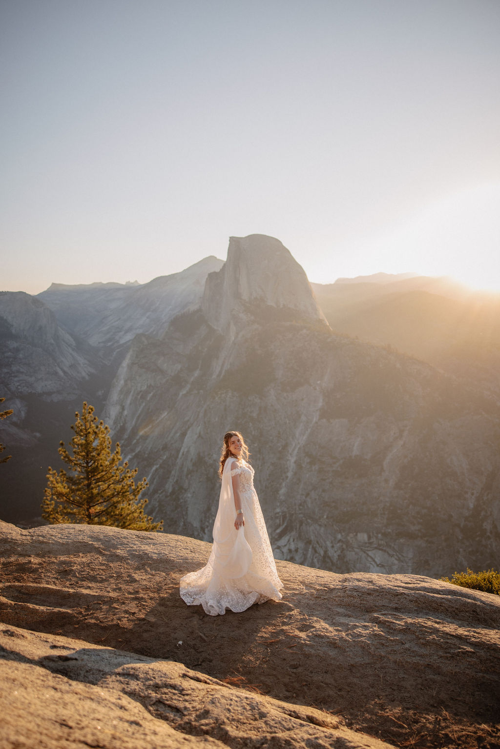 A couple in wedding attire stands on a rocky ledge, overlooking a mountainous landscape at sunrise at an elopement at Yosemite National park in glacier point and mariposa grove