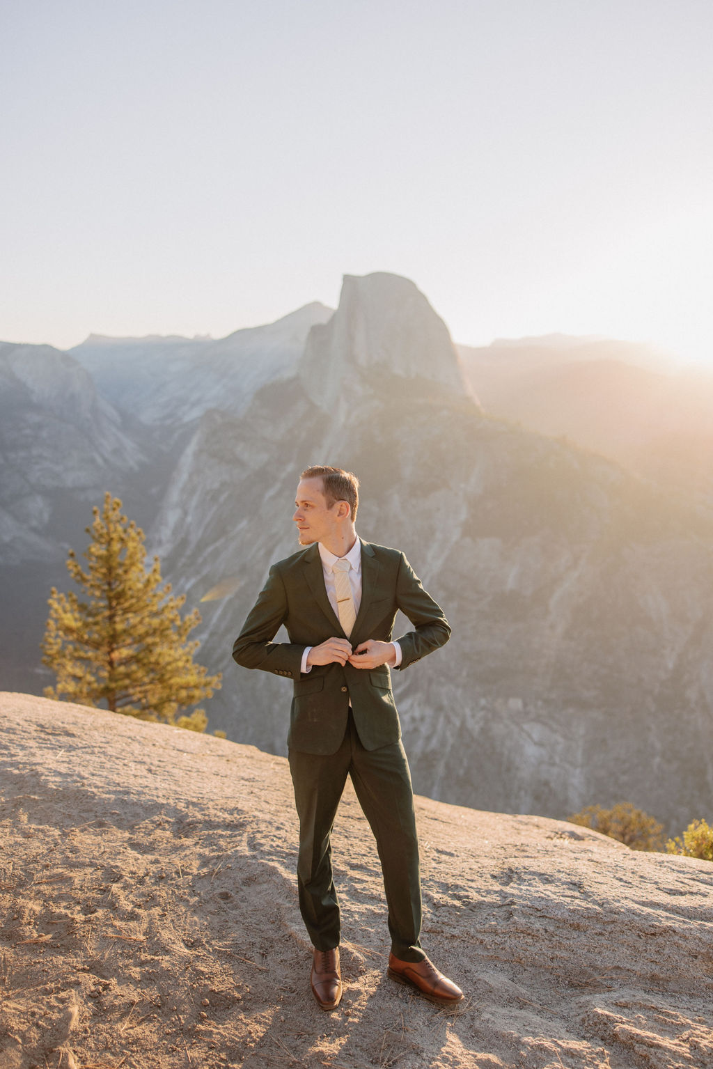 A couple in wedding attire stands on a rocky ledge, overlooking a mountainous landscape at sunrise at an elopement at Yosemite National park in glacier point and mariposa grove