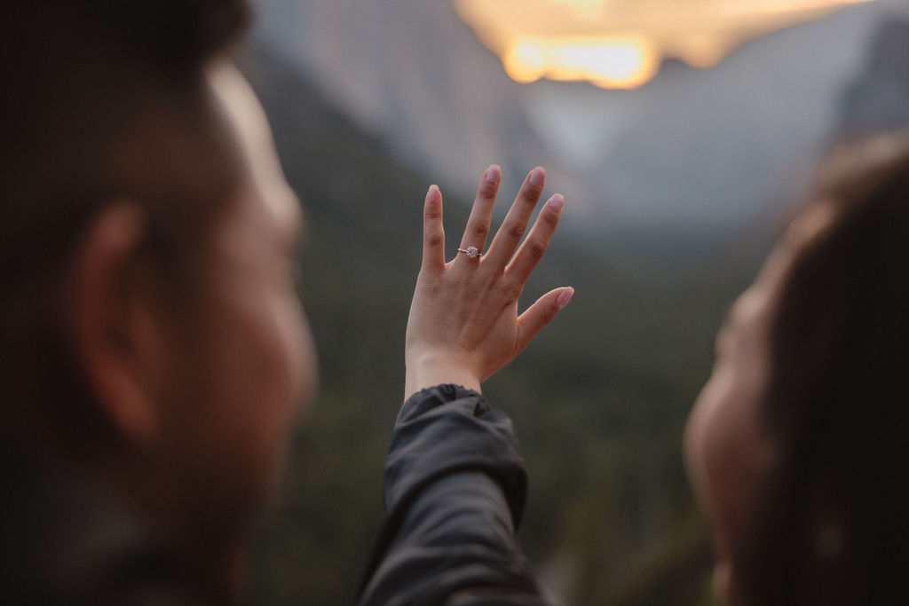 A person holding out a hand with an engagement ring, with blurred view of mountains and sunset in the background.