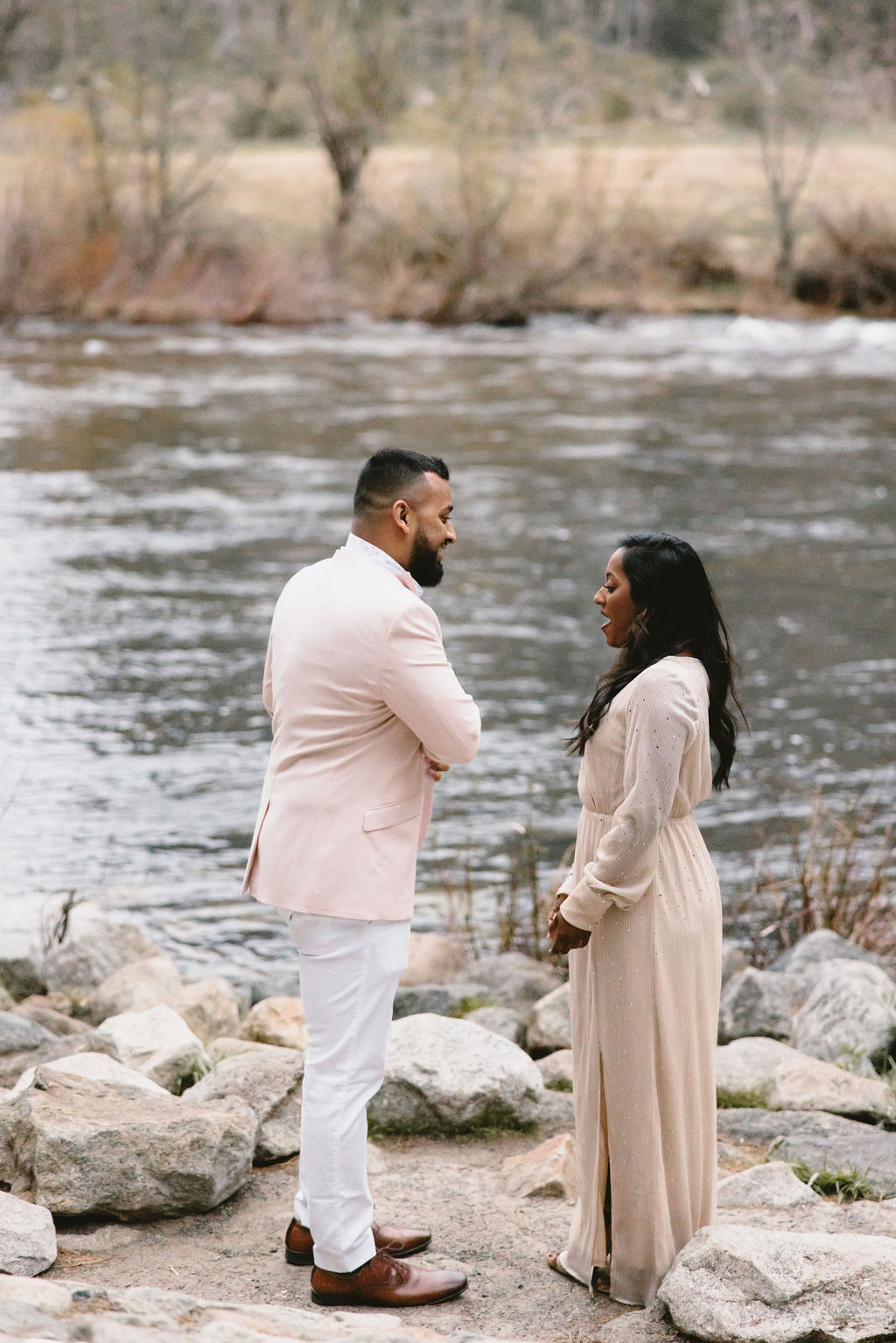 Two people stand facing each other near a riverbank, surrounded by rocks and trees in the background for a surprise proposal at Yosemite