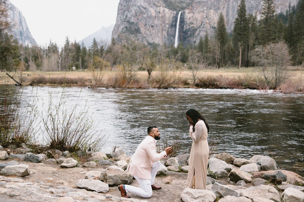 A person kneels and presents a ring to another person by a riverside with mountains and a waterfall in the background.