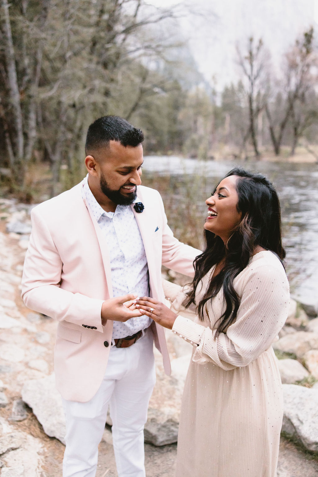 A man and woman are standing by a river in a forested area. Both are smiling and holding hands. The man wears a light pink jacket, and the woman is in a beige dress.