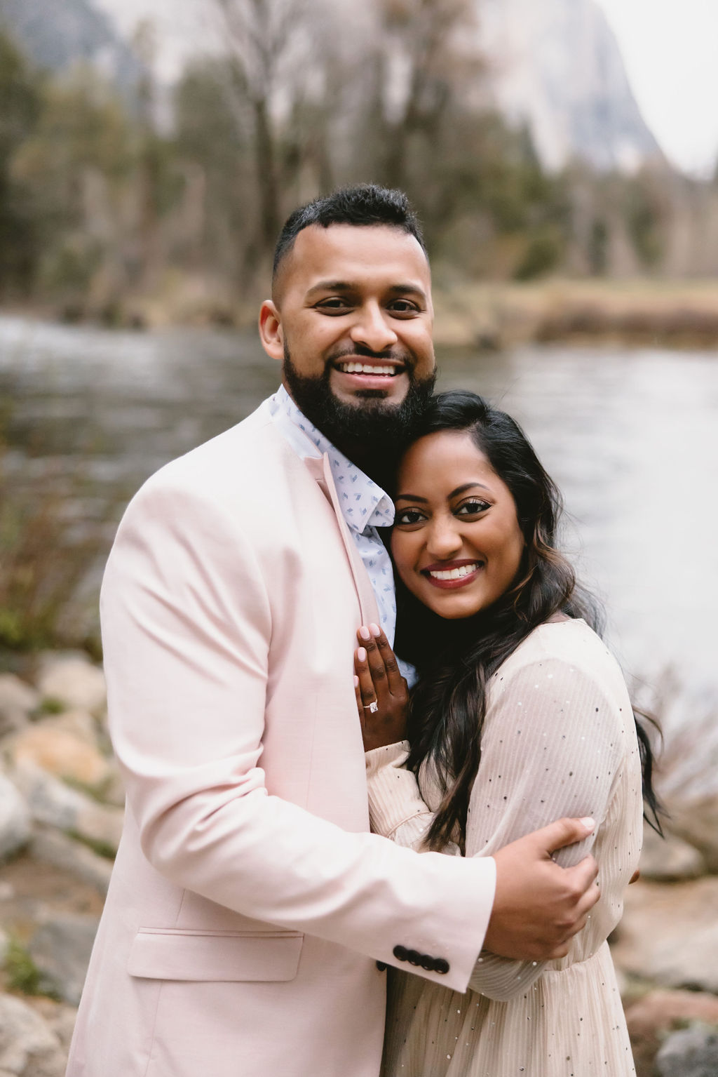 A man and woman are standing by a river in a forested area. Both are smiling and holding hands. The man wears a light pink jacket, and the woman is in a beige dress.