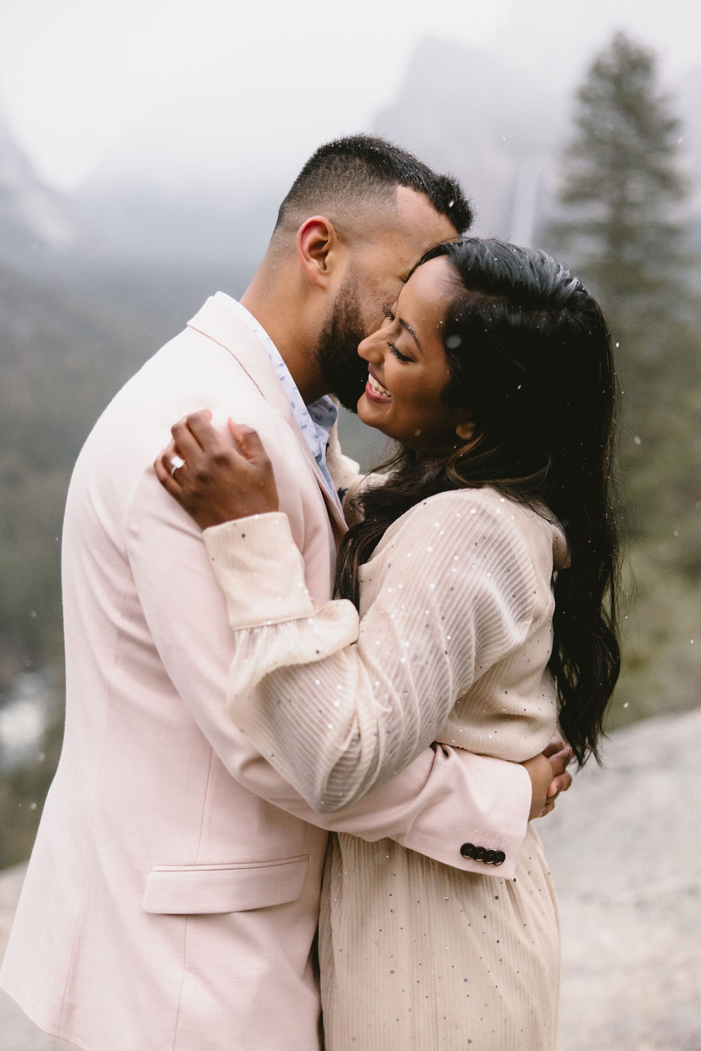 A couple embraces outdoors, wearing light-colored jackets, with a blurred background of trees and mountains.