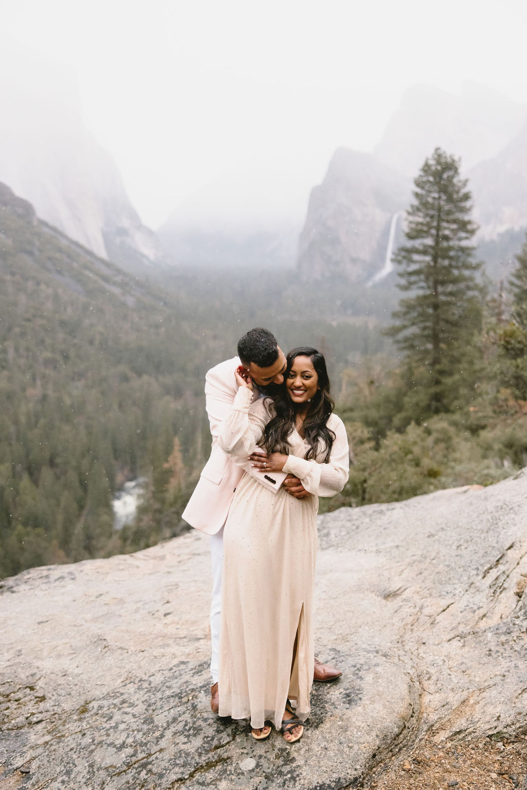 A couple embraces on a rocky ledge overlooking a forested valley with misty mountains in the background.