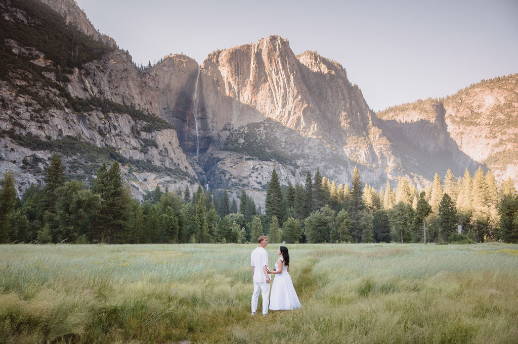 A couple in white attire stands in a grassy meadow with a backdrop of tall mountains and a waterfall.