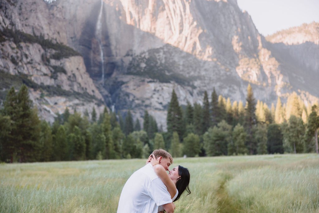 A couple in white attire stands in a grassy meadow with a backdrop of tall mountains and a waterfall.