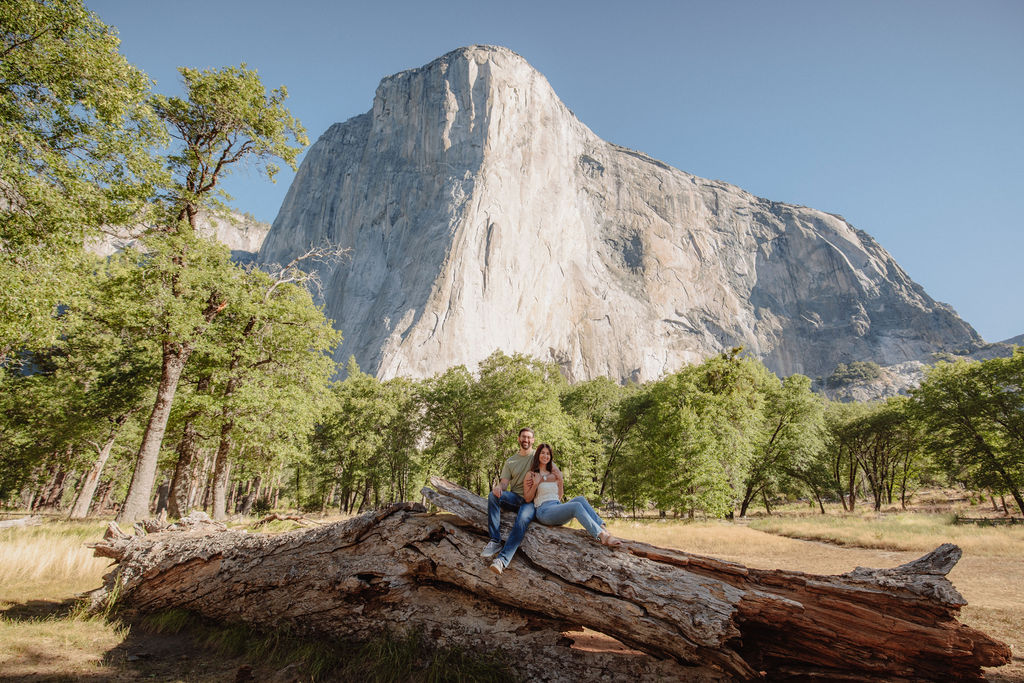 A couple smiling and embracing in front of a rocky river with tall mountains and trees in the background.