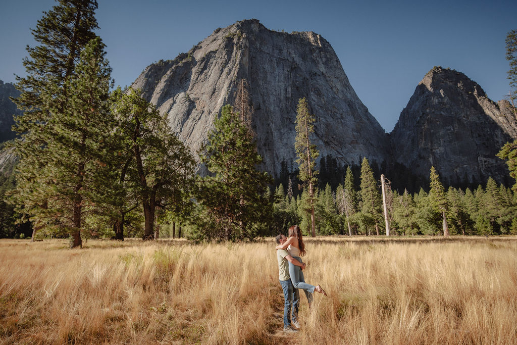 A couple smiling and embracing in front of a rocky river with tall mountains and trees in the background for their yosemite national park engagement photos