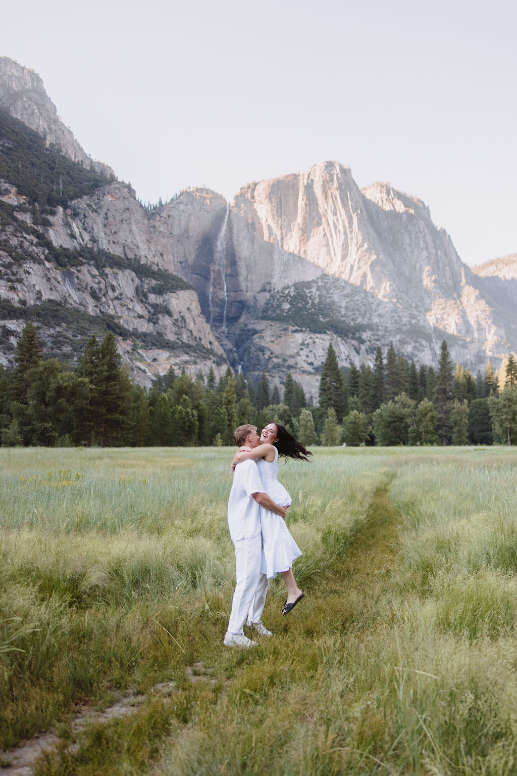 A couple in white attire stands in a grassy meadow with a backdrop of tall mountains and a waterfall.