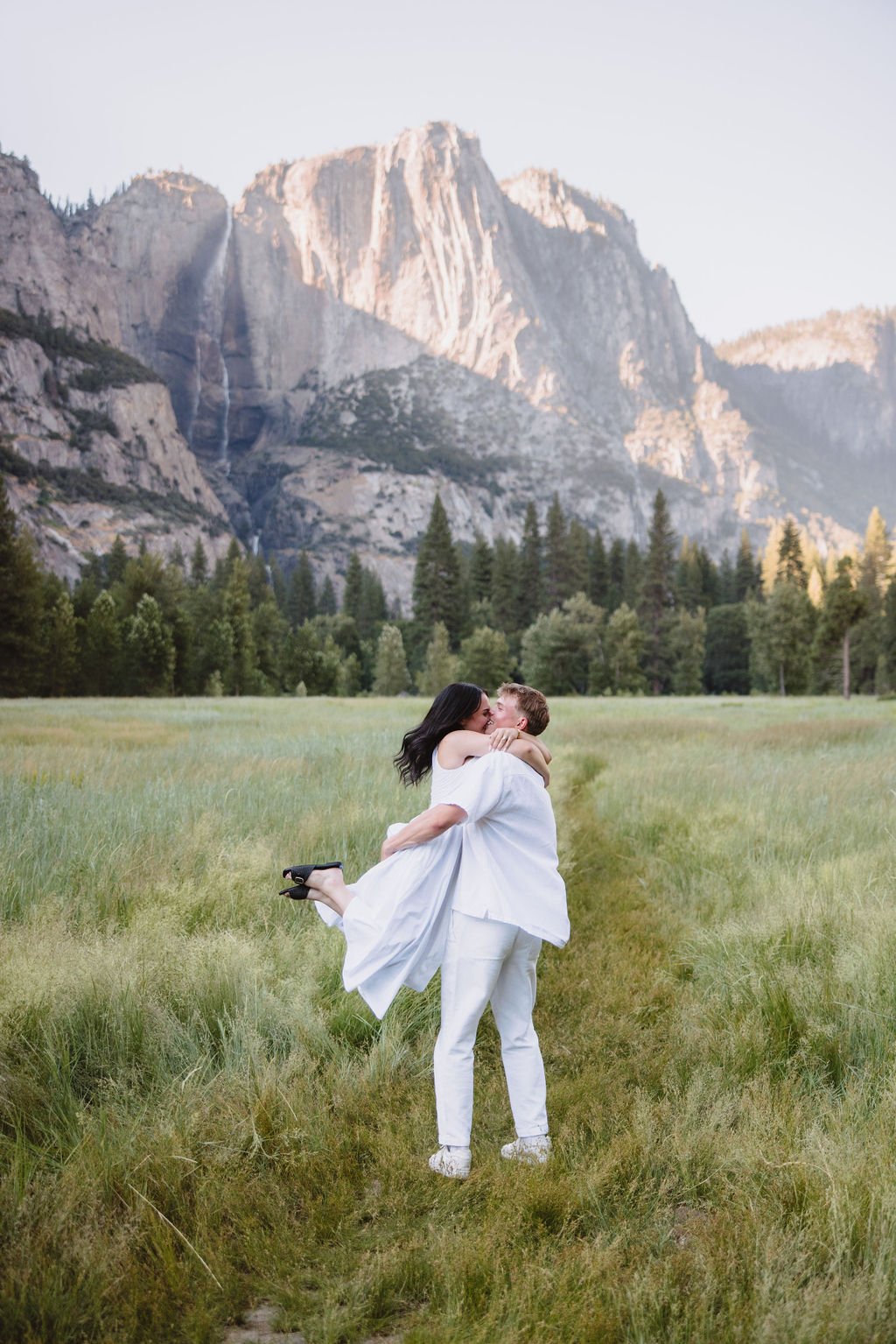 A couple in white attire stands in a grassy meadow with a backdrop of tall mountains and a waterfall.