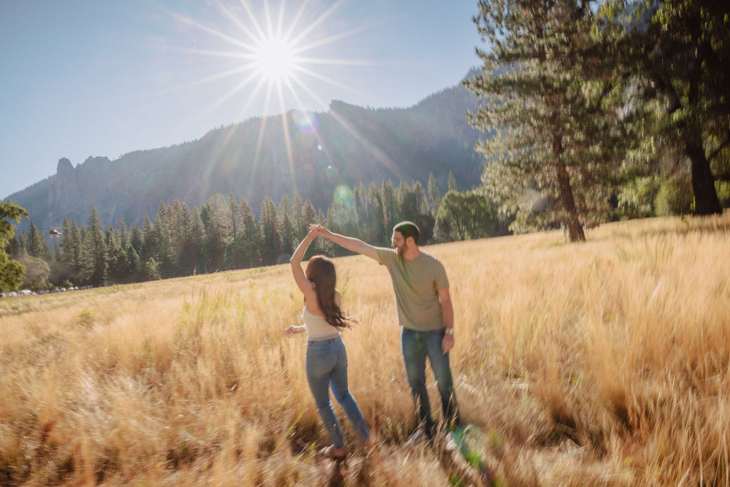 A couple smiling and embracing in front of a rocky river with tall mountains and trees in the background for their yosemite national park engagement photos